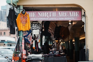 A market stall at The North Arcade showcases vibrant, handmade garments and crafts. A brightly colored sweater dominates the display, with various textiles hanging, including patterned scarves and bags. The atmosphere suggests a bustling marketplace focused on artisanal products.