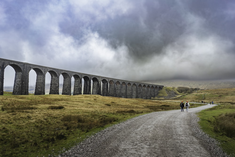 a group of people walking down a dirt road next to a bridge