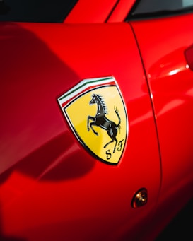 A close-up view of a bright red car featuring the iconic Ferrari logo. The logo features a prancing black horse on a yellow shield background, with the letters 'S' and 'F' at the bottom.