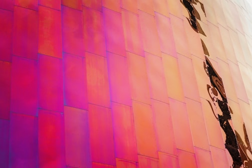 A vibrant wall composed of metallic panels reflects various shades of pink, orange, and purple. The panels are arranged in a staggered pattern, creating an abstract and modern aesthetic. Visible on the right side are darker, swirling reflections.