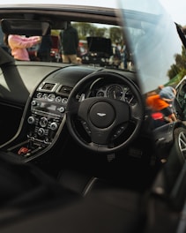A luxury car interior featuring a sleek dashboard with advanced controls, a branded steering wheel, and leather seating. Outside the car, there are people and other vehicles visible, suggesting a car event or gathering.