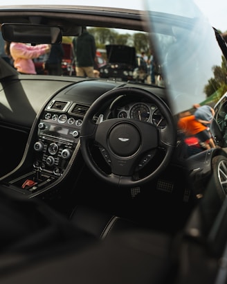 A luxury car interior featuring a sleek dashboard with advanced controls, a branded steering wheel, and leather seating. Outside the car, there are people and other vehicles visible, suggesting a car event or gathering.