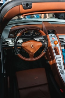 A luxurious car interior featuring a leather steering wheel with the Porsche logo prominently displayed. The dashboard includes analog gauges and an array of buttons, with brown leather accents throughout. The gear shift is a striking wood design, complementing the upscale aesthetics.