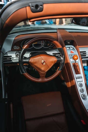 A luxurious car interior featuring a leather steering wheel with the Porsche logo prominently displayed. The dashboard includes analog gauges and an array of buttons, with brown leather accents throughout. The gear shift is a striking wood design, complementing the upscale aesthetics.