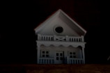 A small, white model house with a gabled roof and a circular window near the top. It has two levels with white railings resembling a porch and balcony.