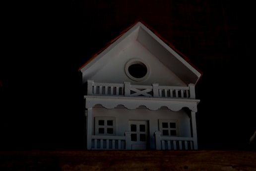 A small, white model house with a gabled roof and a circular window near the top. It has two levels with white railings resembling a porch and balcony.