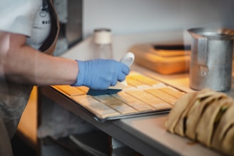 A person wearing a blue glove is using a brush to apply liquid onto rectangular pastry pieces on a baking tray. The setting appears to be a kitchen or bakery, with utensils and a metal pitcher visible.