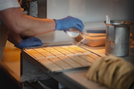 A person wearing blue gloves is sprinkling some type of powder over uniformly arranged rectangular pieces on a tray. The setting appears to be a kitchen or food preparation area, with a metal canister and a stack of items in the foreground.