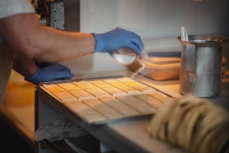 A person wearing blue gloves is sprinkling some type of powder over uniformly arranged rectangular pieces on a tray. The setting appears to be a kitchen or food preparation area, with a metal canister and a stack of items in the foreground.