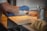 A person wearing blue gloves is sprinkling some type of powder over uniformly arranged rectangular pieces on a tray. The setting appears to be a kitchen or food preparation area, with a metal canister and a stack of items in the foreground.