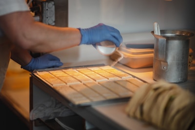 A person wearing blue gloves is sprinkling some type of powder over uniformly arranged rectangular pieces on a tray. The setting appears to be a kitchen or food preparation area, with a metal canister and a stack of items in the foreground.