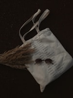 A canvas tote bag with a list of grocery items printed on it, featuring categories like bread, milk, cookies, and other essentials. The bag is laid on a dark surface with a bundle of dried grass partially covering it. A pair of round sunglasses is placed on the bag.