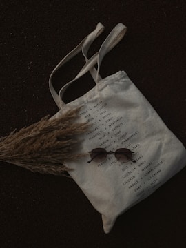 A canvas tote bag with a list of grocery items printed on it, featuring categories like bread, milk, cookies, and other essentials. The bag is laid on a dark surface with a bundle of dried grass partially covering it. A pair of round sunglasses is placed on the bag.