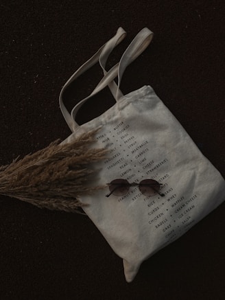 A canvas tote bag with a list of grocery items printed on it, featuring categories like bread, milk, cookies, and other essentials. The bag is laid on a dark surface with a bundle of dried grass partially covering it. A pair of round sunglasses is placed on the bag.