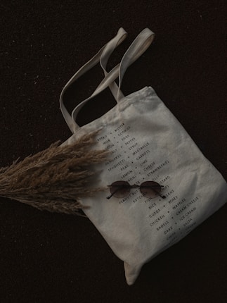 A canvas tote bag with a list of grocery items printed on it, featuring categories like bread, milk, cookies, and other essentials. The bag is laid on a dark surface with a bundle of dried grass partially covering it. A pair of round sunglasses is placed on the bag.