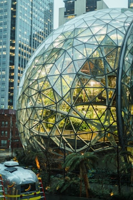 A large glass geodesic dome with a lush interior of plants and trees is set amidst high-rise buildings. The structure features a complex network of triangular glass facets with vibrant greenery visible inside. In front of the dome, there is a silver vehicle, possibly an airstream, parked with decals on it. The surrounding architecture and modern urban setting contribute to an innovative and futuristic ambiance.