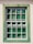 A window with a grid-like frame is set in a light-colored wall. The frame is painted in a weathered green, providing a rustic appearance. The glass panes have a reflective quality, showing hints of greenery from the surrounding environment.