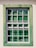A window with a grid-like frame is set in a light-colored wall. The frame is painted in a weathered green, providing a rustic appearance. The glass panes have a reflective quality, showing hints of greenery from the surrounding environment.