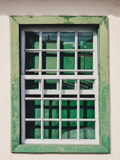 A window with a grid-like frame is set in a light-colored wall. The frame is painted in a weathered green, providing a rustic appearance. The glass panes have a reflective quality, showing hints of greenery from the surrounding environment.