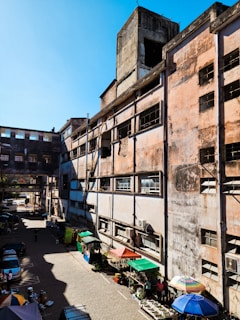 A large, weathered industrial building with multiple stories and a faded exterior. The structure features rectangular windows, some with broken panes, and is surrounded by a small market area with various stalls. The street level includes colorful umbrellas and market tables, with potted plants and other goods. A few vehicles and people are visible in the narrow alleyway.