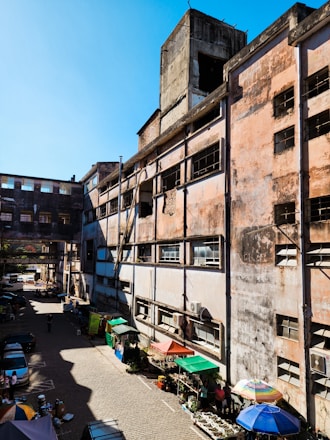 A large, weathered industrial building with multiple stories and a faded exterior. The structure features rectangular windows, some with broken panes, and is surrounded by a small market area with various stalls. The street level includes colorful umbrellas and market tables, with potted plants and other goods. A few vehicles and people are visible in the narrow alleyway.