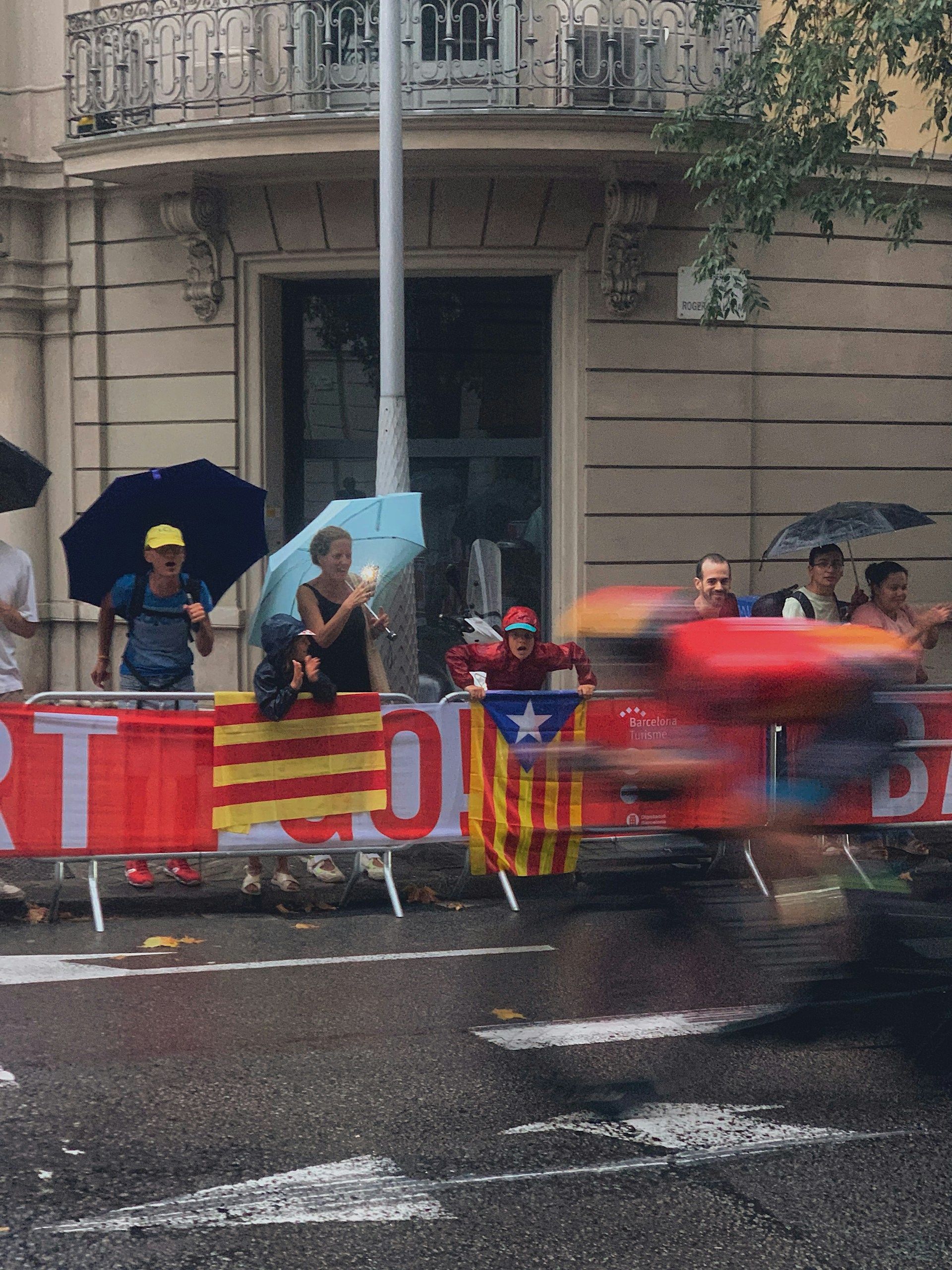 A group of people standing under umbrellas beside a street are watching a cycling event. Red and yellow Catalan flags are draped along the barrier, and a cyclist in red attire is speeding past, appearing blurred due to motion.