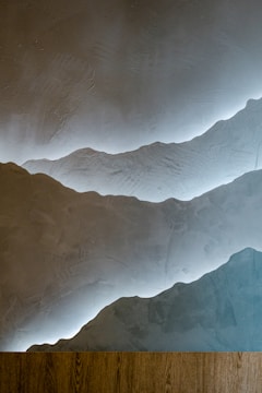 A textured wall with a layered design resembling mountains, highlighted by soft lighting from behind the layers. At the bottom, there is a wooden plank or surface, creating a contrast with the smooth plaster-like texture of the wall.