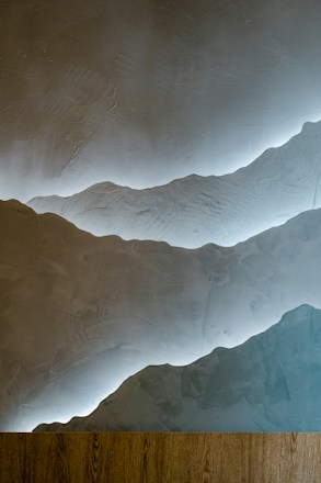A textured wall with a layered design resembling mountains, highlighted by soft lighting from behind the layers. At the bottom, there is a wooden plank or surface, creating a contrast with the smooth plaster-like texture of the wall.