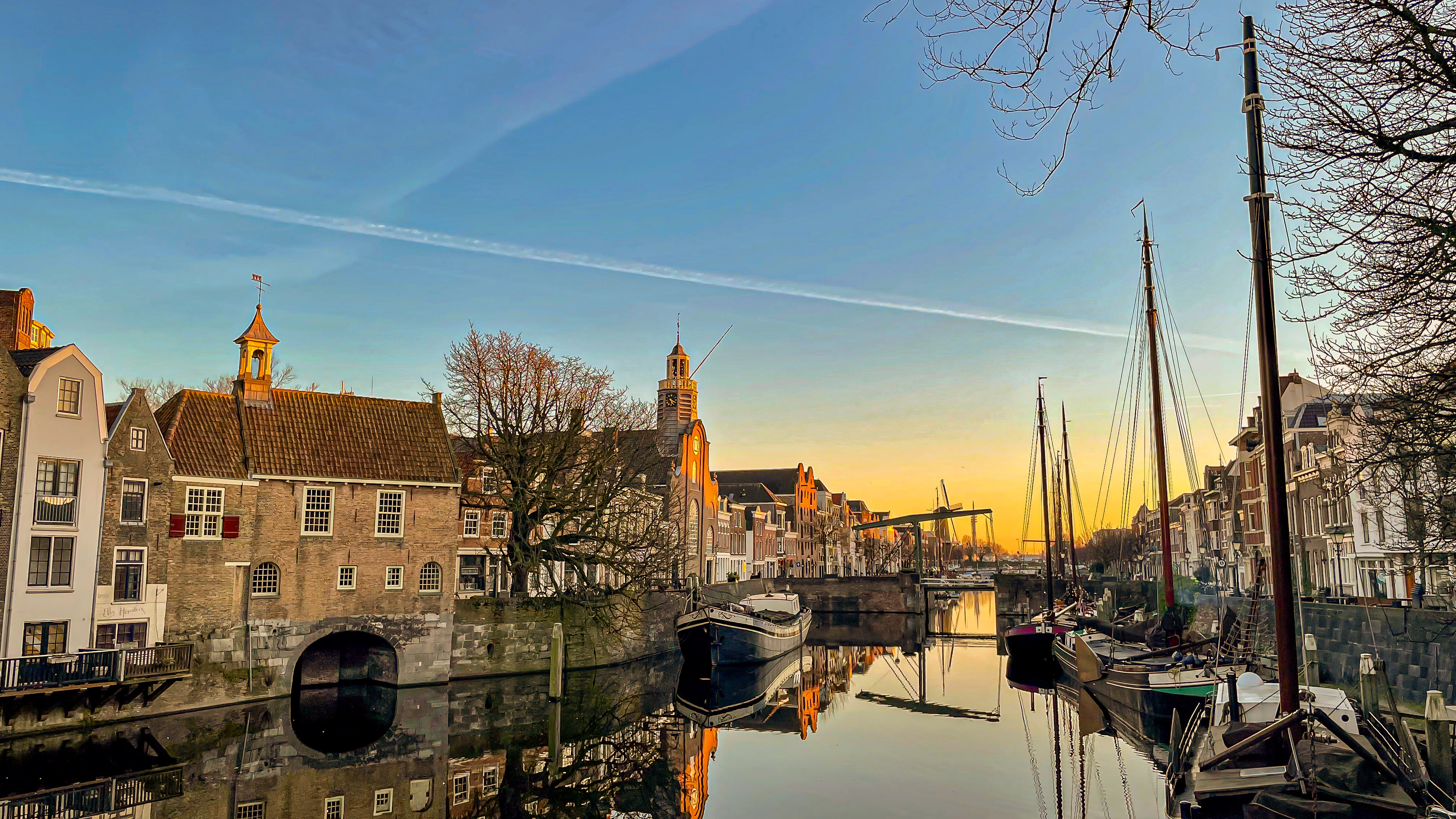 Delfshaven's old harbour in Rotterdam at low winter sun, bare trees and gabled brick houses glowing gold along a mirror-still canal with masted barges moored on the right.