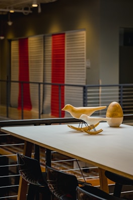 A modern indoor setting features a table with decorative items, including two abstract bird sculptures and an egg-shaped object. The background showcases a wall with vertical panels in red and white colors, and a railing runs along the mezzanine level.
