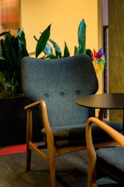 A cozy room featuring a mid-century modern armchair with wooden armrests and a dark upholstered cushion. In the background, there are lush green plants, and a small round table is positioned next to the chair. The lighting is warm and inviting, adding to the comfortable atmosphere.