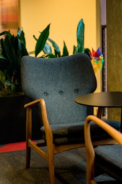 A cozy room featuring a mid-century modern armchair with wooden armrests and a dark upholstered cushion. In the background, there are lush green plants, and a small round table is positioned next to the chair. The lighting is warm and inviting, adding to the comfortable atmosphere.