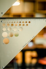 A metal triangular ruler with a series of circular holes of varying sizes is positioned against a blurred background. The ruler is marked with inch measurements along its edges and is labeled with the brand 'FARIGATE' and specifications 'by STAMP INC., 45°-90°'. The background is softly lit, with a geometric design, suggesting an indoor setting with subdued colors.
