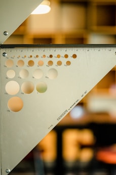 A metal triangular ruler with a series of circular holes of varying sizes is positioned against a blurred background. The ruler is marked with inch measurements along its edges and is labeled with the brand 'FARIGATE' and specifications 'by STAMP INC., 45°-90°'. The background is softly lit, with a geometric design, suggesting an indoor setting with subdued colors.