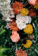 A vibrant floral arrangement featuring a variety of flowers, including white roses, orange chrysanthemums, bright yellow pom-pom flowers, and lush green foliage. The composition is dense with blooms and leaves, creating a lush and colorful display.