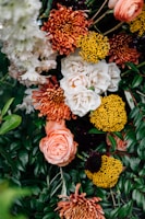 A vibrant floral arrangement featuring a variety of flowers, including white roses, orange chrysanthemums, bright yellow pom-pom flowers, and lush green foliage. The composition is dense with blooms and leaves, creating a lush and colorful display.