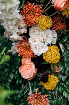 A vibrant floral arrangement featuring a variety of flowers, including white roses, orange chrysanthemums, bright yellow pom-pom flowers, and lush green foliage. The composition is dense with blooms and leaves, creating a lush and colorful display.