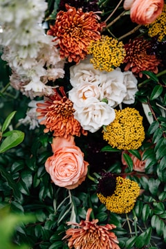 A vibrant floral arrangement featuring a variety of flowers, including white roses, orange chrysanthemums, bright yellow pom-pom flowers, and lush green foliage. The composition is dense with blooms and leaves, creating a lush and colorful display.