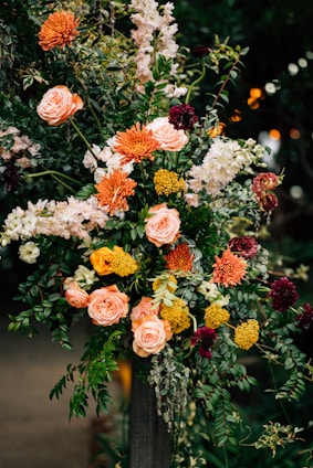 A vibrant floral arrangement featuring a variety of blossoms including roses, marigolds, and other colorful flowers set against a backdrop of lush green foliage. The arrangement is composed of warm colors like orange, yellow, peach, and burgundy, with white flowers that add contrast. The display is elegantly crafted, showcasing the natural beauty and diversity of the flowers.
