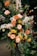 A vibrant floral arrangement featuring a variety of blossoms including roses, marigolds, and other colorful flowers set against a backdrop of lush green foliage. The arrangement is composed of warm colors like orange, yellow, peach, and burgundy, with white flowers that add contrast. The display is elegantly crafted, showcasing the natural beauty and diversity of the flowers.
