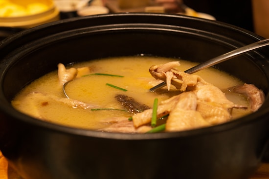 A steaming bowl of soup with chunks of chicken and pieces of green onion in a dark, round pot. The broth appears thick and creamy. A metal spoon rests in the pot, poised to scoop up the contents.