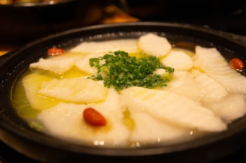 Thinly sliced pieces of white fish arranged in a circular pattern around the edge of a black bowl filled with a yellowish broth. Green chopped scallions are sprinkled in the center, adding a pop of color. Red Goji berries are scattered among the fish slices.