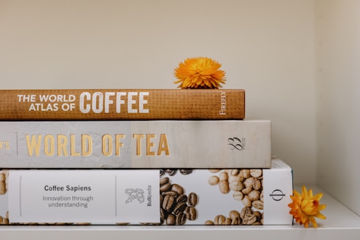 A stack of three books is neatly arranged, each with a distinct subject on coffee and tea. The books are titled 'The World Atlas of Coffee,' 'World of Tea,' and 'Coffee Sapiens.' Two bright orange flowers are placed decoratively around the books, adding a touch of natural beauty to the scene.