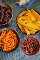 An arrangement of five bowls containing different snacks: red grapes in a wooden bowl, potato chips in a blue bowl, sliced mangoes in a brown bowl, cheese flavored snacks in a dark brown bowl, and a mix of nuts in a red bowl.