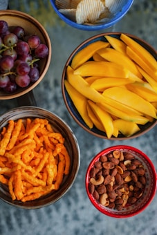 An arrangement of five bowls containing different snacks: red grapes in a wooden bowl, potato chips in a blue bowl, sliced mangoes in a brown bowl, cheese flavored snacks in a dark brown bowl, and a mix of nuts in a red bowl.