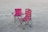 Two pink folding camping chairs are positioned on a gravelly surface. The chairs have armrests and cup holders, with one slightly more reclined than the other. The scene lacks any people, and the chairs appear well-used.
