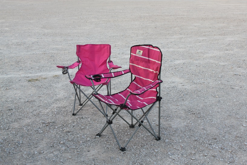 Two pink folding camping chairs are positioned on a gravelly surface. The chairs have armrests and cup holders, with one slightly more reclined than the other. The scene lacks any people, and the chairs appear well-used.