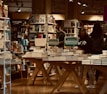 A bookstore filled with shelves of books and organized tables displaying stacks of books. Several people are browsing, some wearing casual clothes. Promotional signs are visible, advertising a special offer for double stamps on purchases. The lighting gives a warm and cozy atmosphere, with wooden elements adding to the inviting setting.