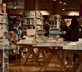 A bookstore filled with shelves of books and organized tables displaying stacks of books. Several people are browsing, some wearing casual clothes. Promotional signs are visible, advertising a special offer for double stamps on purchases. The lighting gives a warm and cozy atmosphere, with wooden elements adding to the inviting setting.