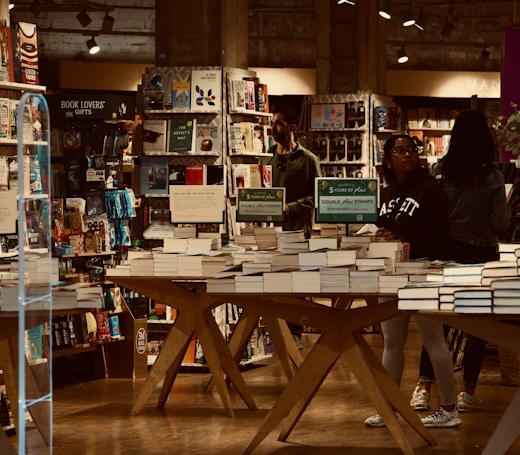 A bookstore filled with shelves of books and organized tables displaying stacks of books. Several people are browsing, some wearing casual clothes. Promotional signs are visible, advertising a special offer for double stamps on purchases. The lighting gives a warm and cozy atmosphere, with wooden elements adding to the inviting setting.